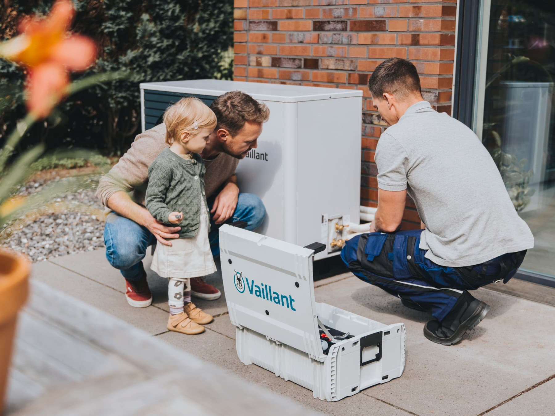 Papa und Kind stehen neben einer Wärmepumpe, während Techniker an dem Gerät arbeitet
